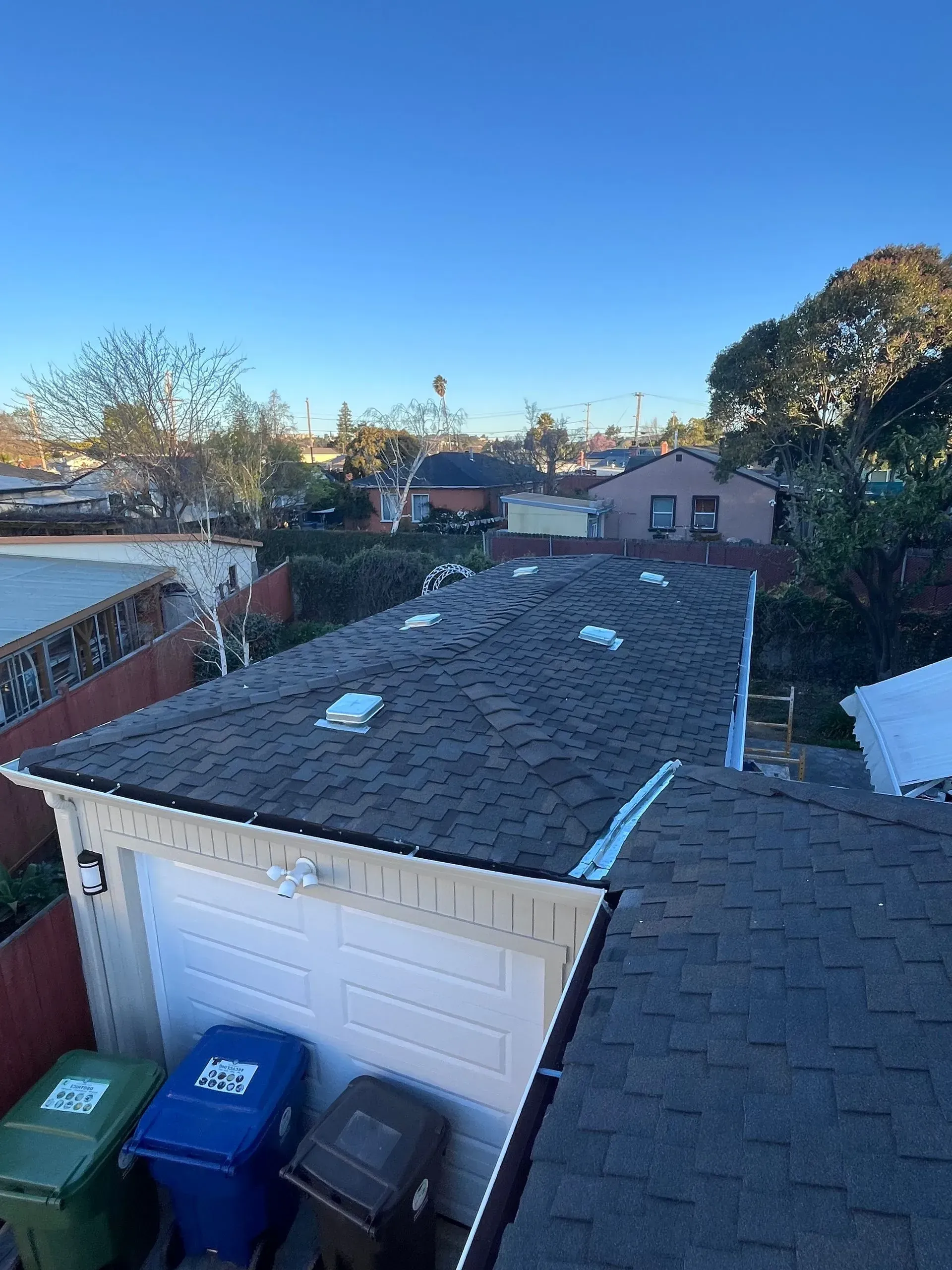 View of a dark shingled roof with several vents, a garage, trash bins, and a residential neighborhood under a blue sky.