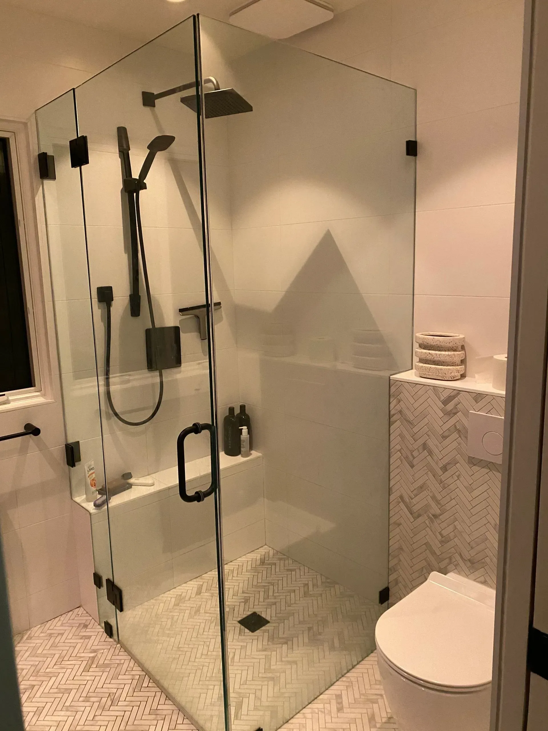 Glass-walled shower with black fixtures and built-in bench. White walls and herringbone tile floor in modern bathroom.