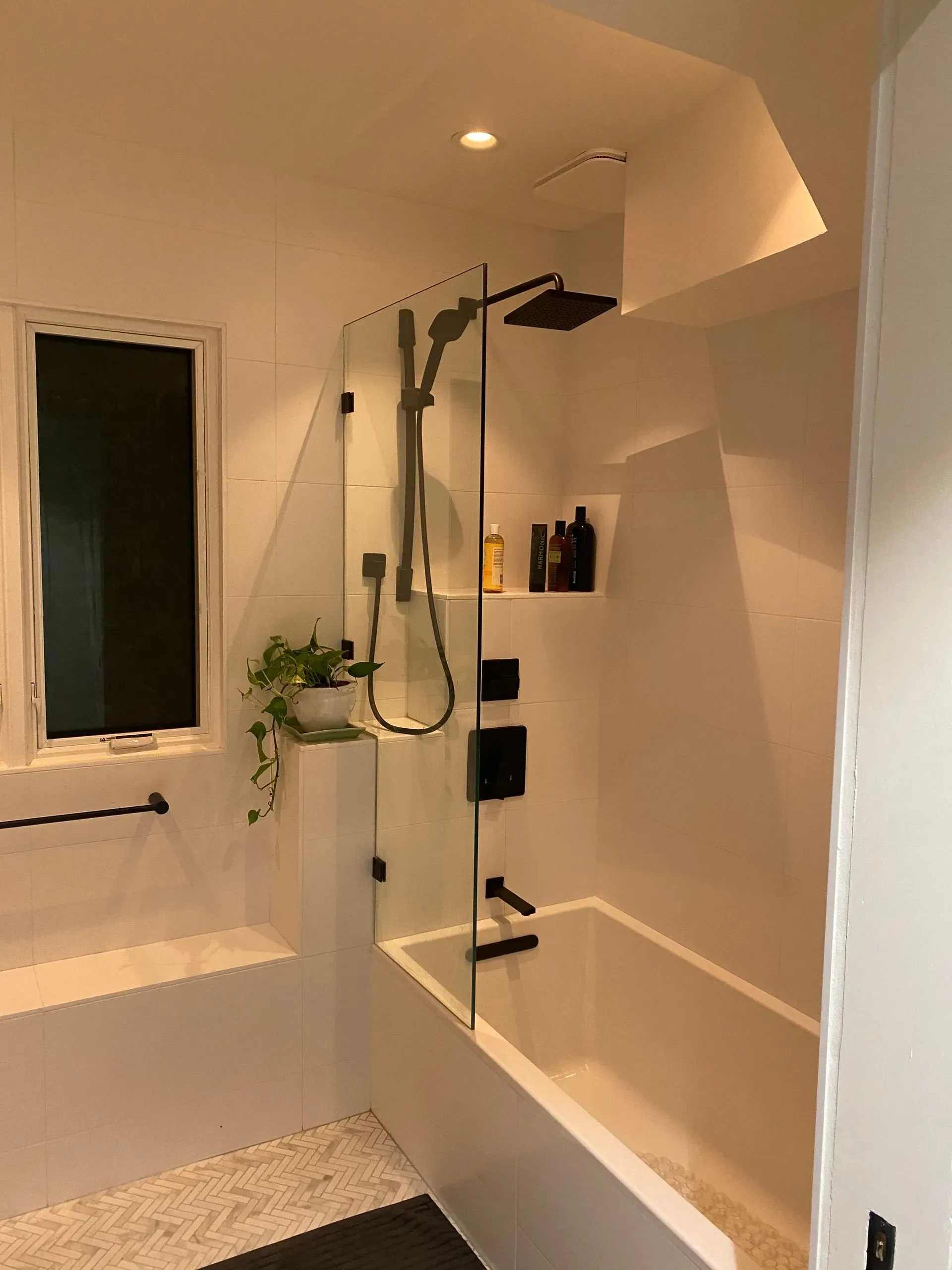 Modern white bathroom with a glass shower enclosure over a white tub. Black fixtures and a small window are visible.