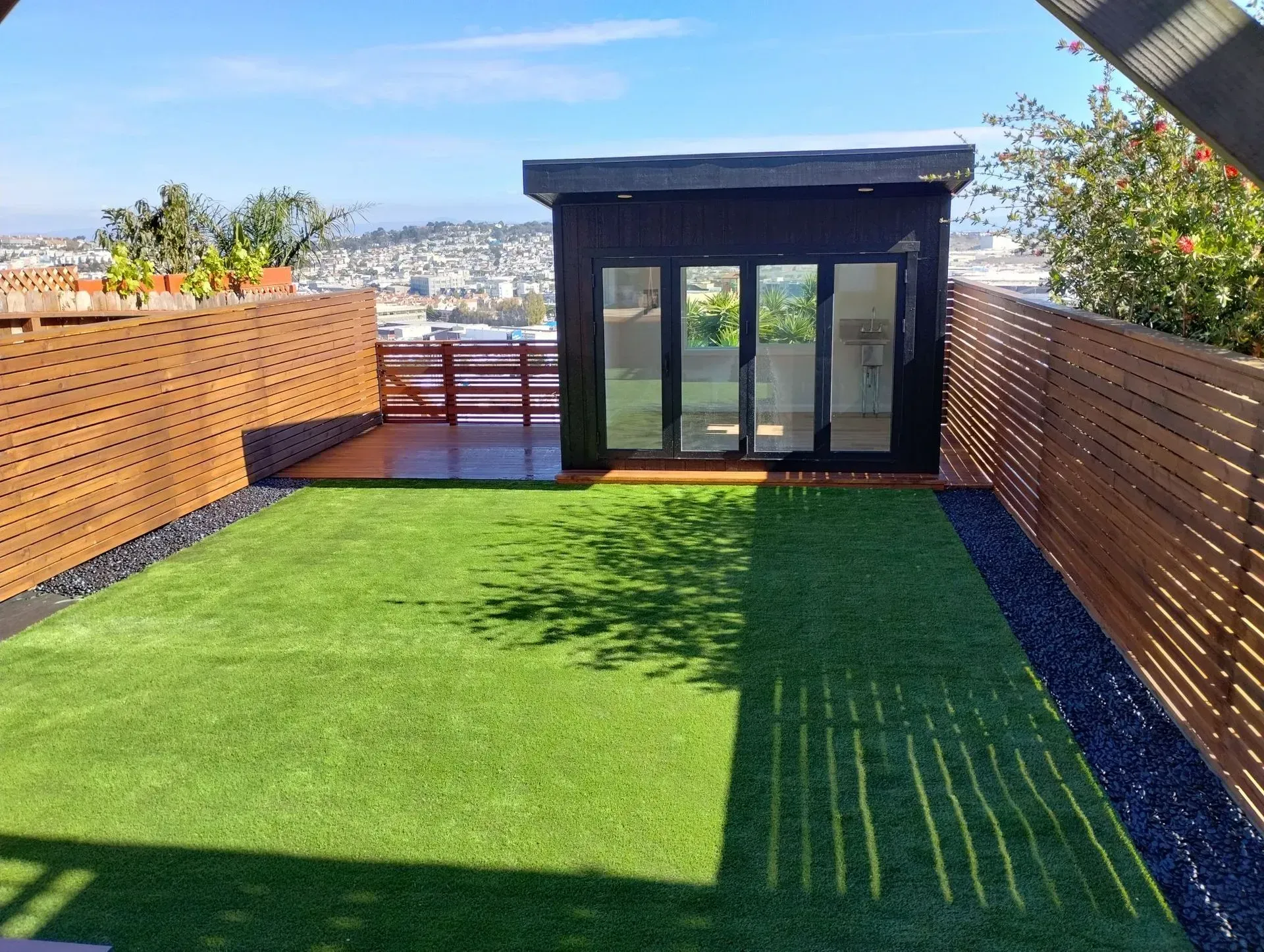 Backyard with artificial grass, wooden fence, modern shed with glass doors, overlooking a city.