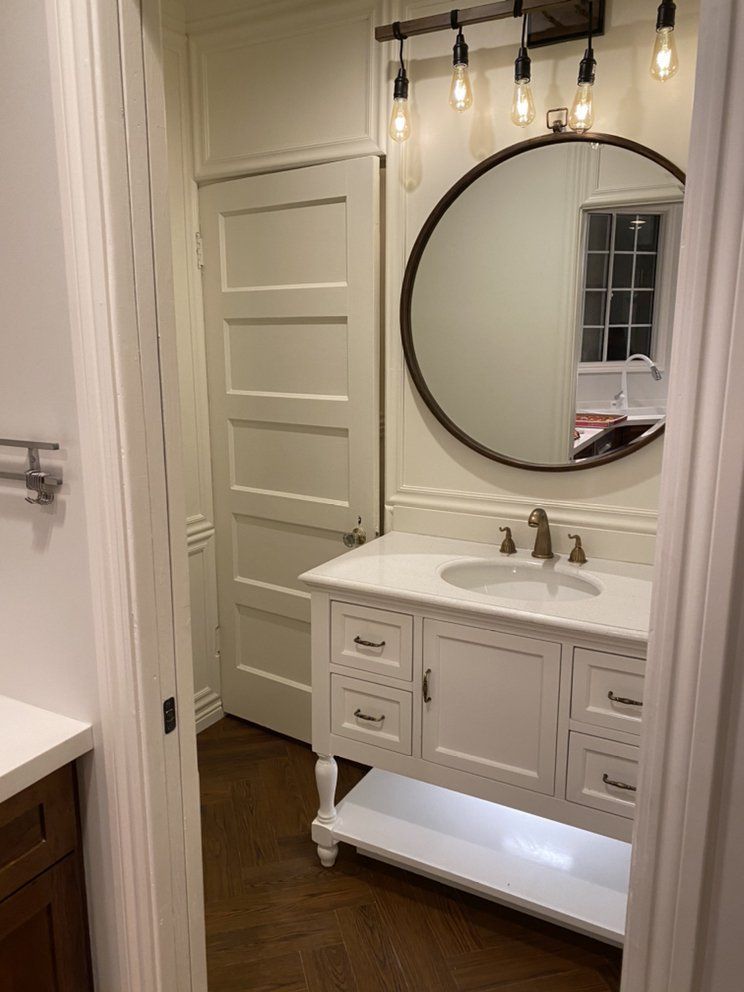 Bathroom with a white vanity, round mirror, and overhead lighting. The door is visible.