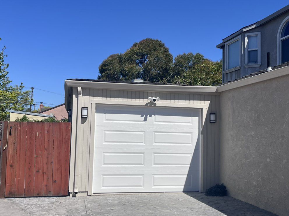 White garage door with lights, next to a brown fence and beige wall, under a blue sky.