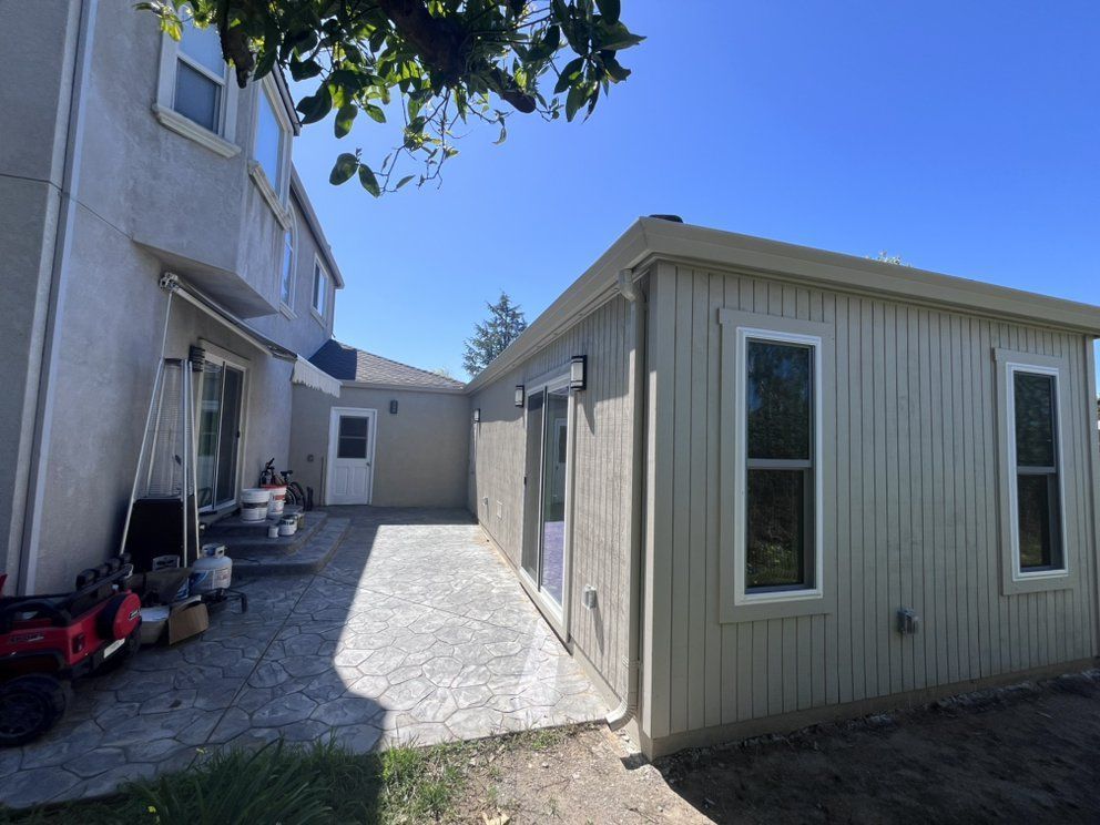 Backyard with a two-story beige house and a light tan single-story building; concrete pathway connects them.