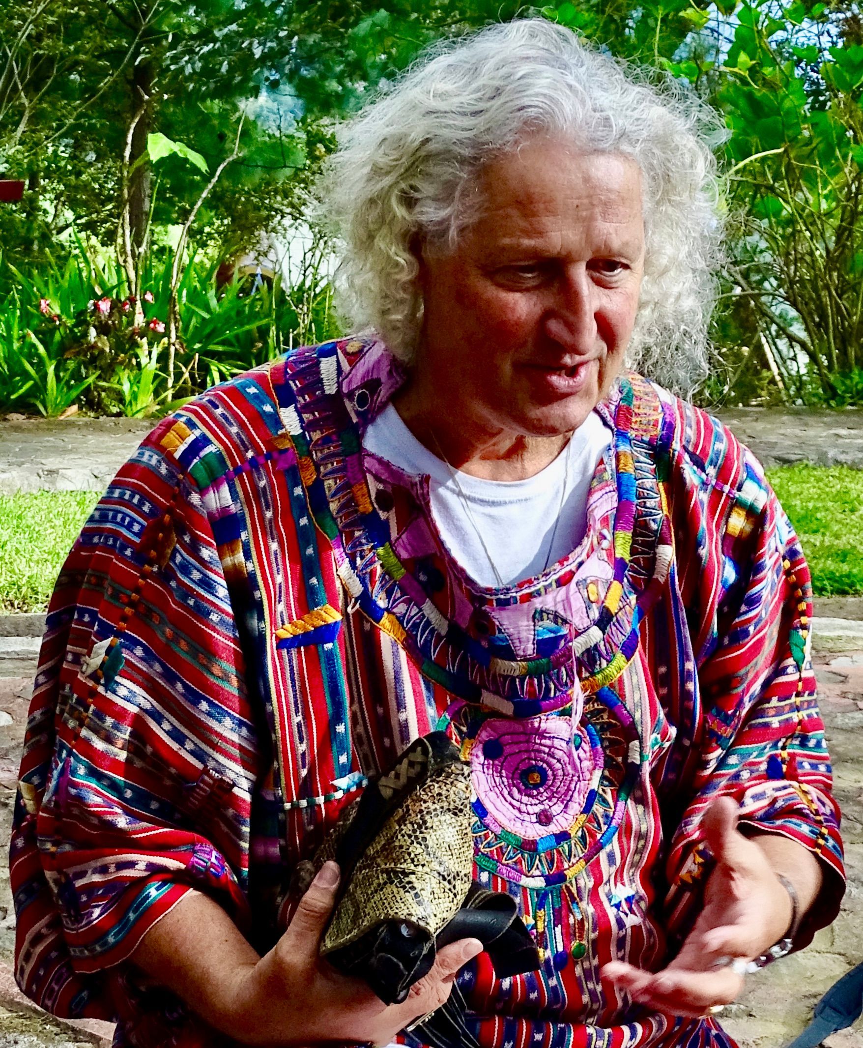 Mann mit lockigem, weißem Haar, der ein buntes, gemustertes Hemd trägt, hält im Freien einen Gegenstand.