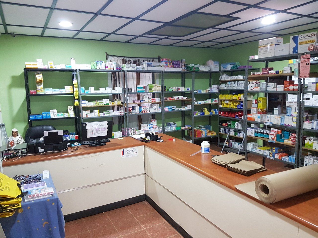 Innenansicht einer Apotheke mit Medikamentenregalen hinter einem Tresen. Grüne Wände, weiße Decke, braune Arbeitsplatten.