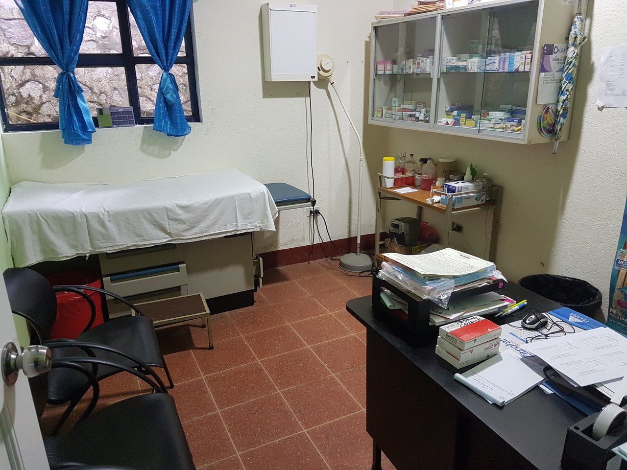 Untersuchungsraum mit Untersuchungsliege, Schreibtisch, Stühlen und Schränken voller medizinischer Instrumente.