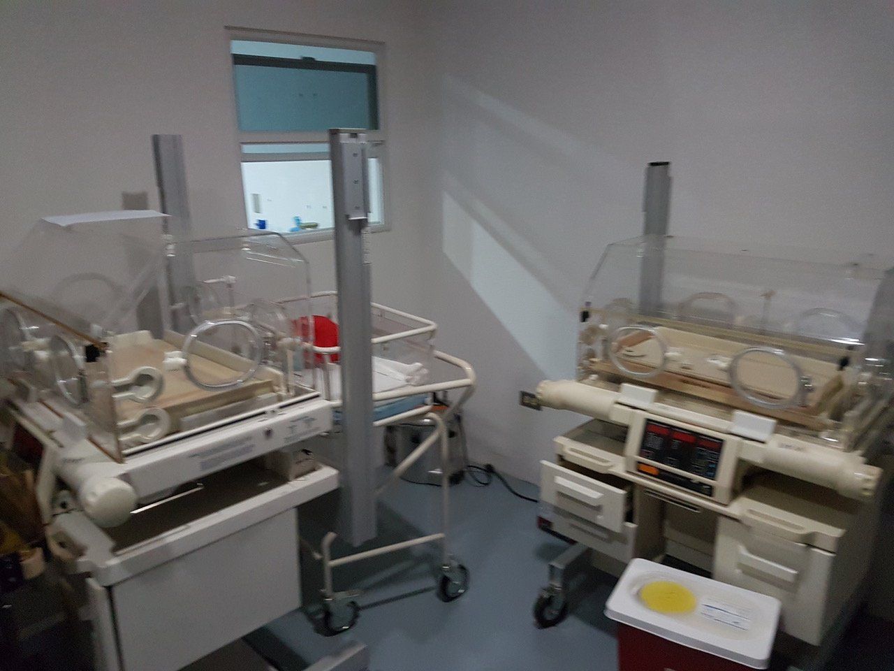Zwei Neugeborenen-Inkubatoren in einem Krankenzimmer, mit medizinischer Ausrüstung und einem kleinen Fenster.