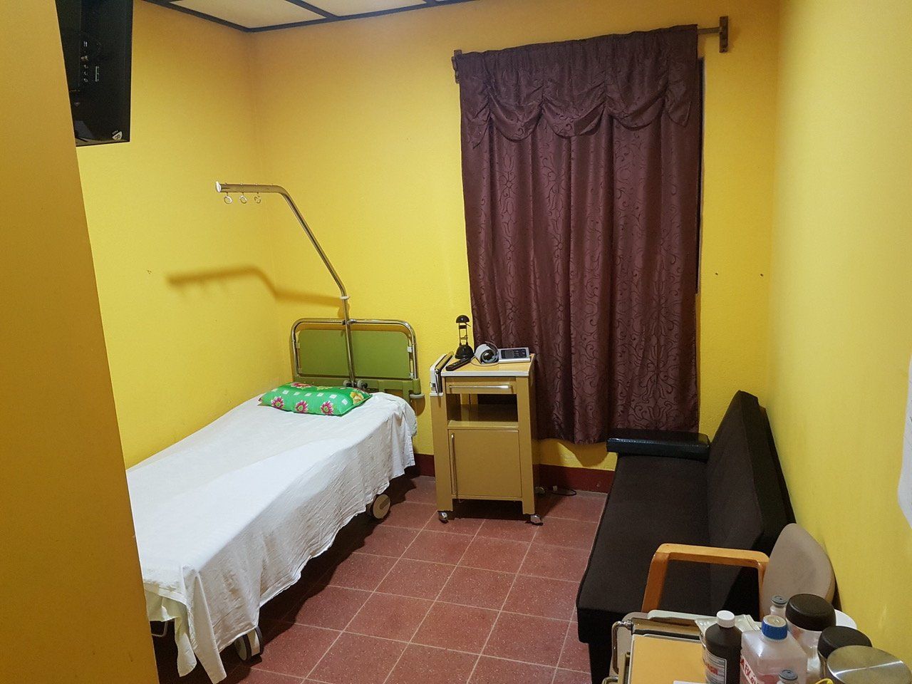 Ein kleines, gelb getünchtes Behandlungszimmer mit einem Bett, einem kleinen Schrank und einem Sofa.