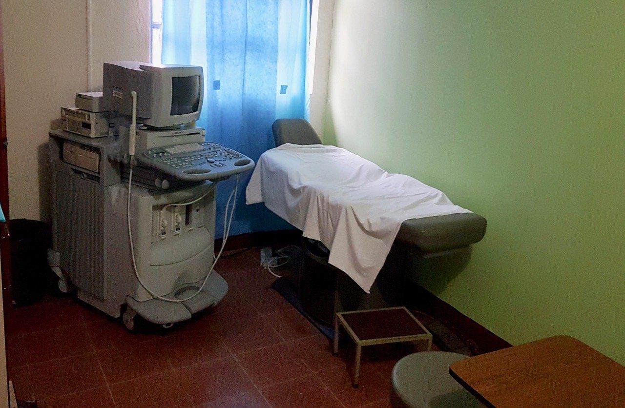 Ein Ultraschallgerät in einem Untersuchungsraum mit einer Liege und einem kleinen Tisch.