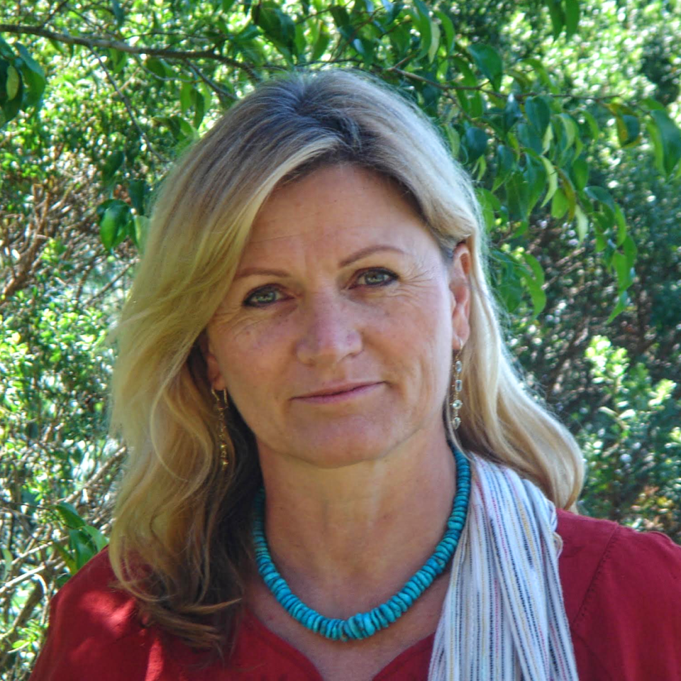 Frau mit blonden Haaren, die im Freien lächelt, trägt ein rotes Hemd, eine türkisfarbene Halskette und einen weißen Schal.