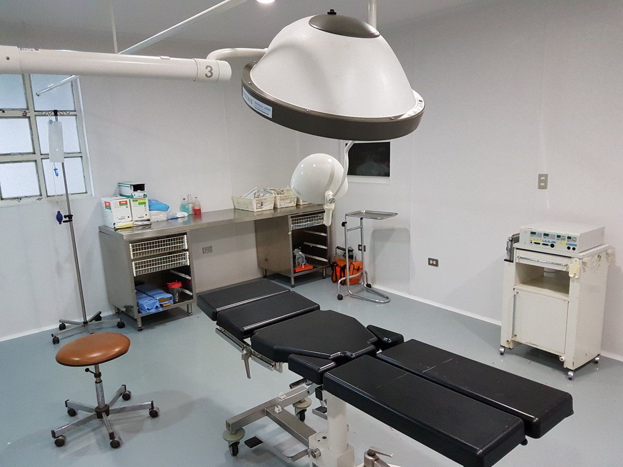Operationssaal mit OP-Leuchten, OP-Tisch, medizinischen Geräten und einem Hocker.