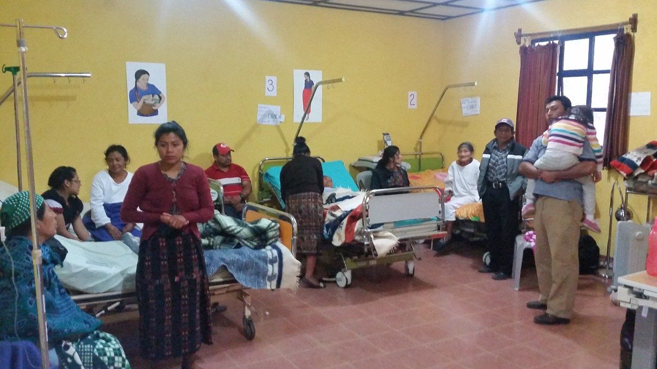 Krankenzimmer mit Patienten in Betten, Angehörige anwesend. Gelbe Wände, verschiedene medizinische Geräte sichtbar.