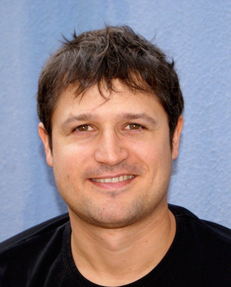 Mann mit dunklen Haaren lächelt in die Kamera, trägt ein schwarzes Hemd und steht vor blauem Hintergrund.