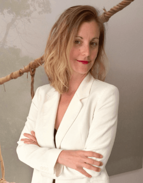 Frau in einem weißen Blazer, die Arme verschränkt, schaut in die Kamera.