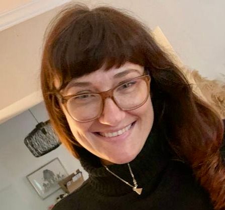 Frau mit braunen Haaren und Brille lächelt; sie trägt einen schwarzen Rollkragenpullover und eine Halskette.