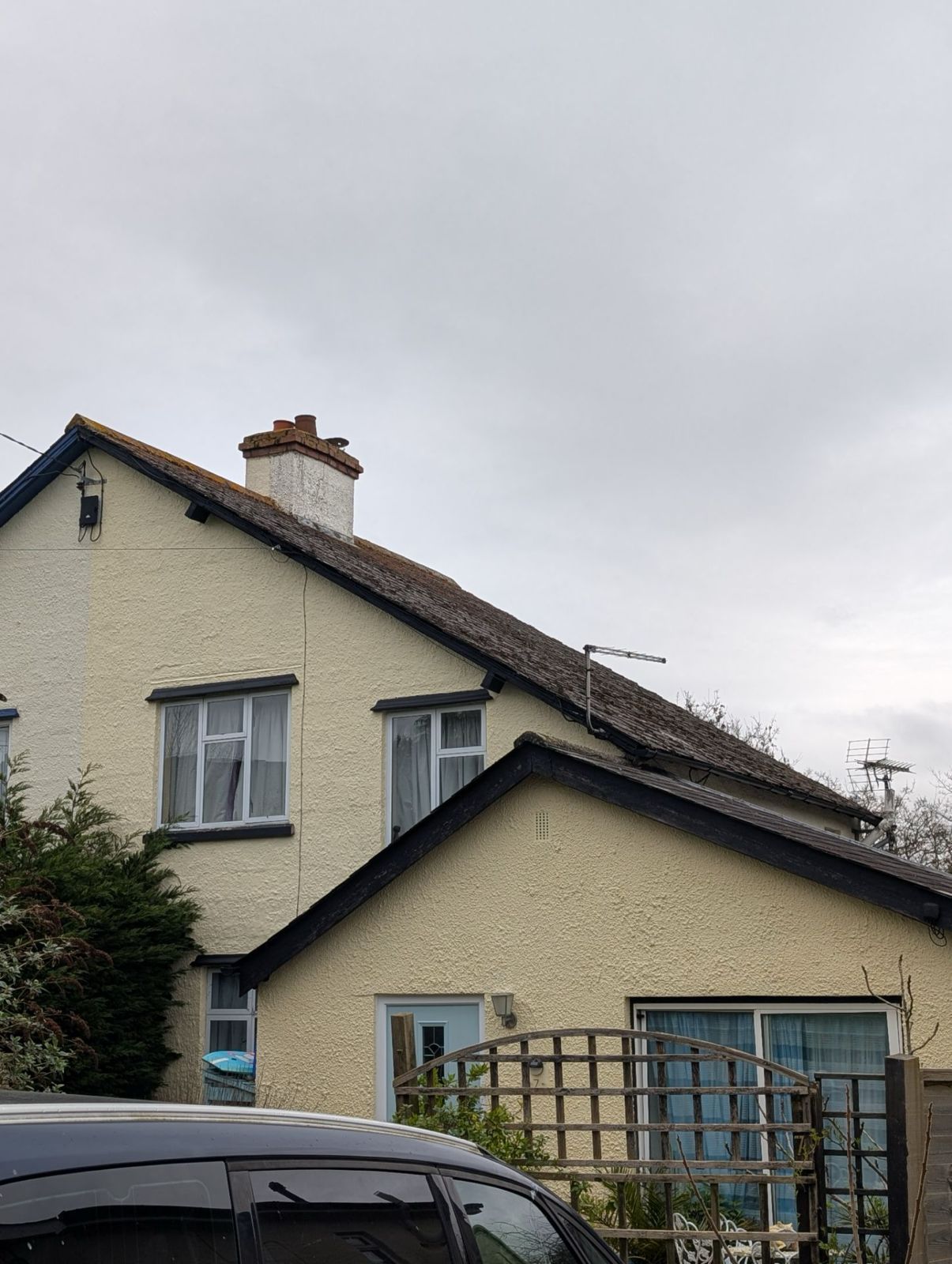 Small cream-colored house with a sloped roof and front trellis under a cloudy sky, with a car in front