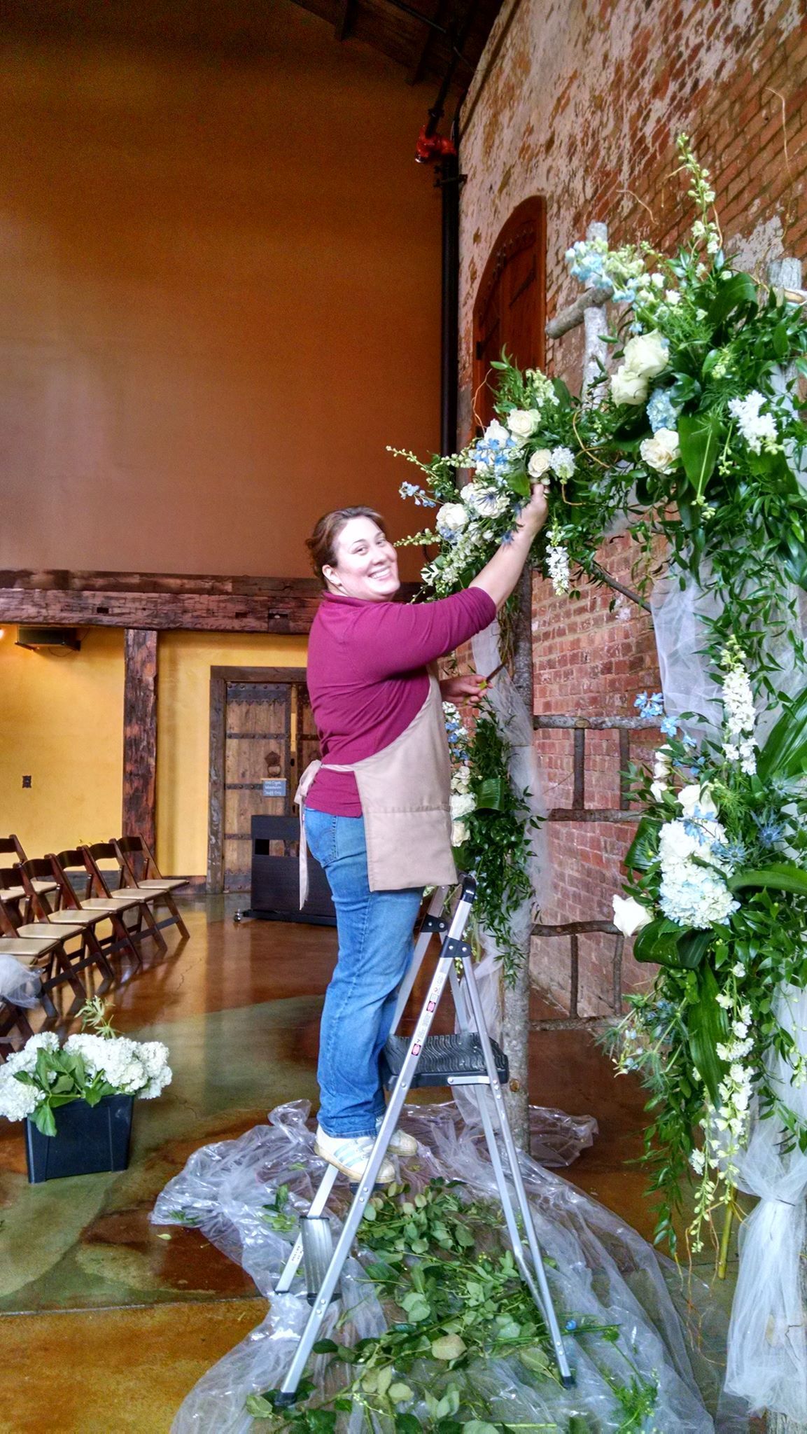 Woman arranging flowers on an archway in a venue, wearing a maroon shirt and jeans.