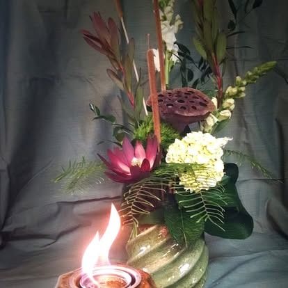 Floral arrangement with a lit candle, featuring pink water lily and green foliage.