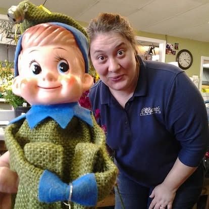 Woman smiles next to a vintage elf doll inside a flower shop.