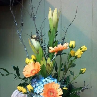 Floral arrangement with orange gerbera daisies, yellow roses, blue hydrangeas, and tall green foliage.