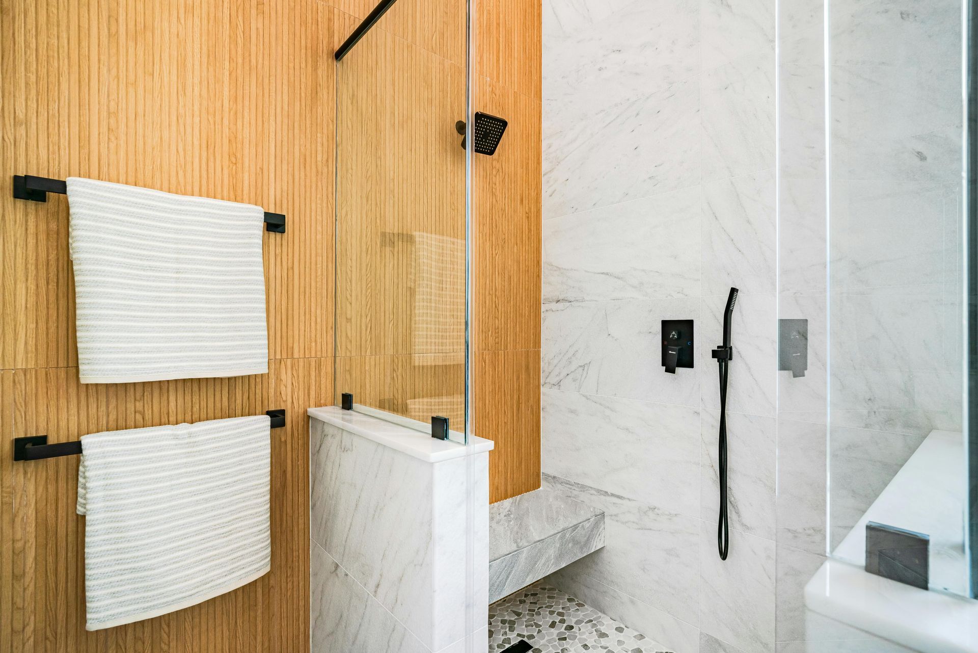 Modern bathroom with wood accent wall, glass shower door, and white marble accents. Black fixtures and white towels.