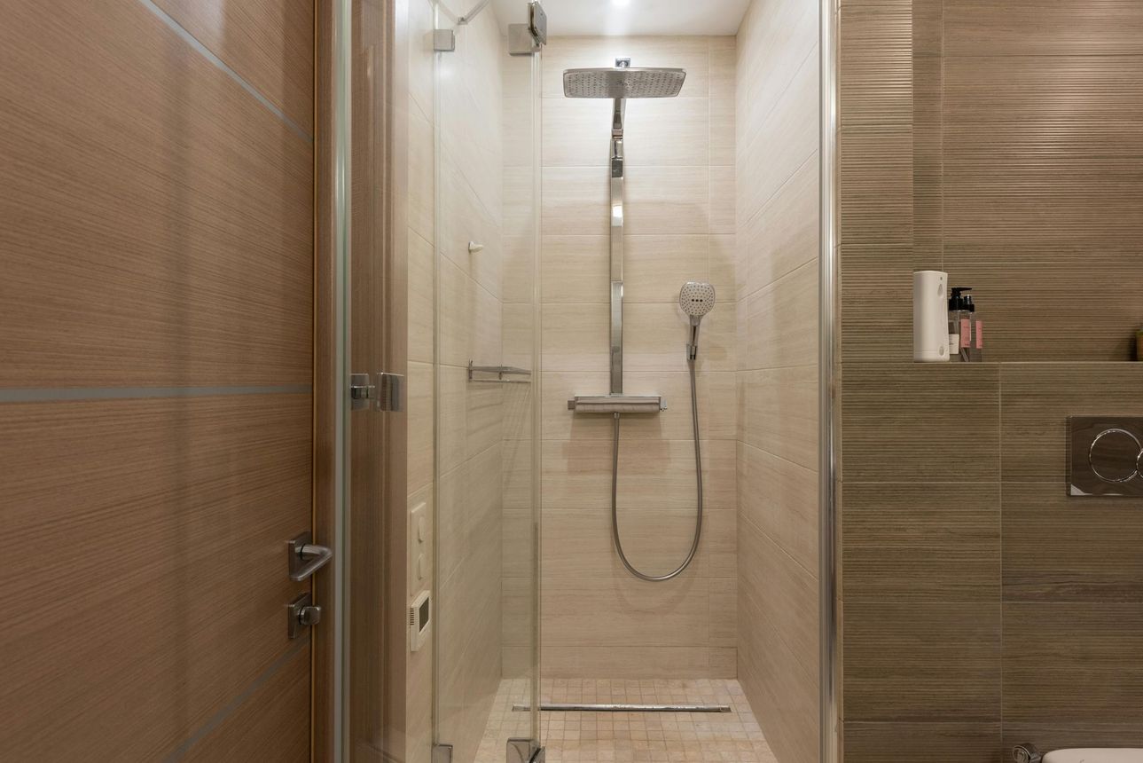 Shower stall with glass doors and a rainfall showerhead. Beige tile and wood door are visible.