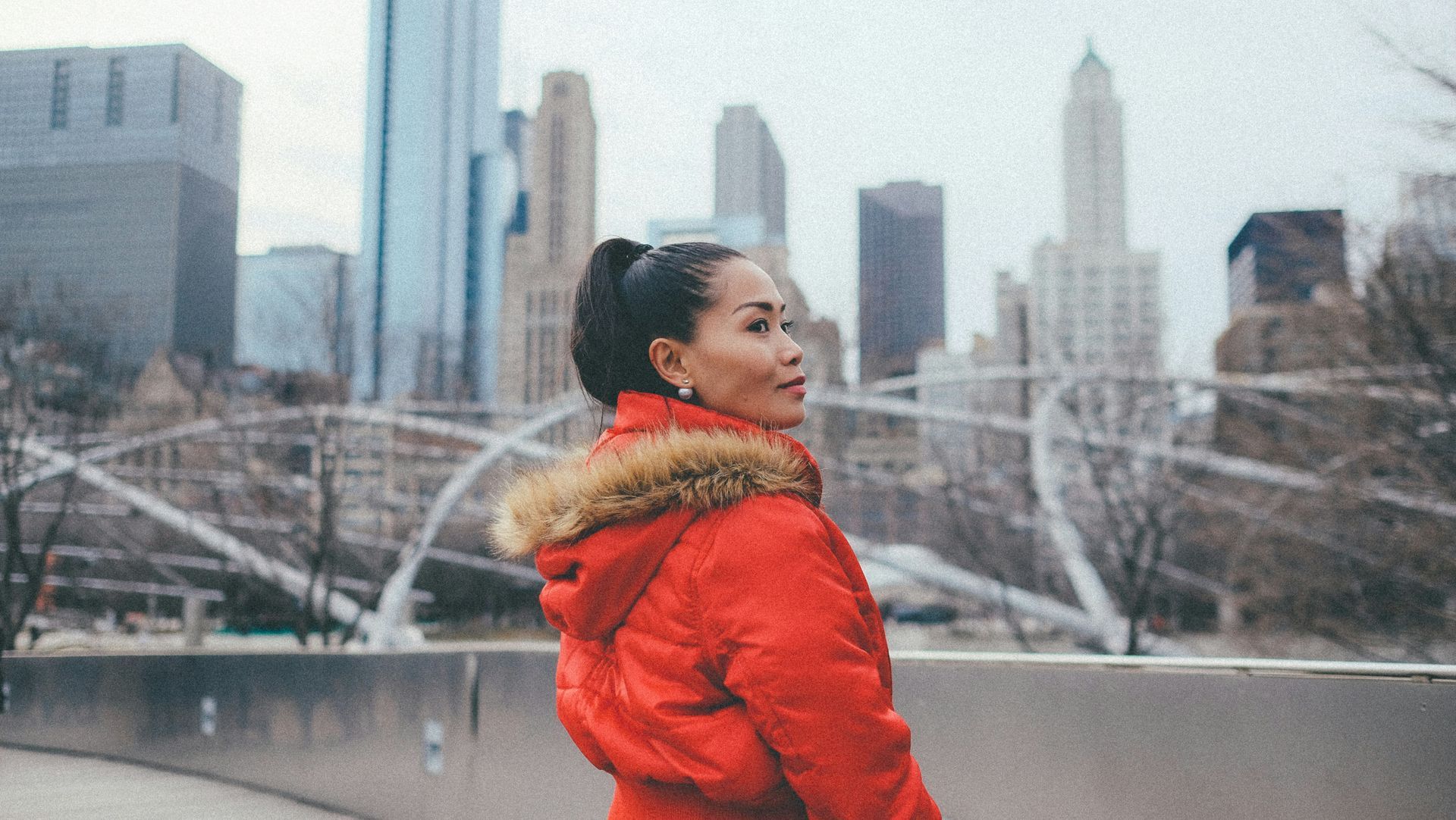 A woman in a red jacket with a fur hood is standing on a bridge in front of a city skyline.