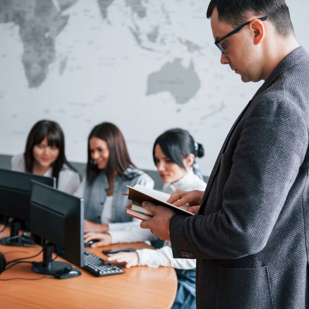 Man in glasses and suit jacket, reading book near three women working at computers.