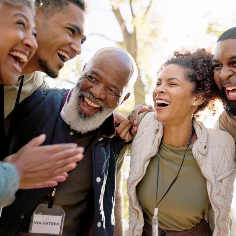 Group of diverse volunteers laughing, outdoors; an older man with a gray beard is at the center.