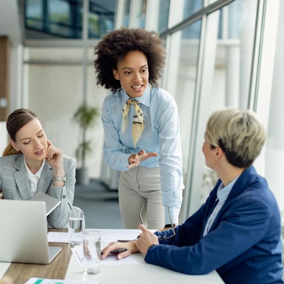 Three women in an office meeting: one speaking, gesturing; others listening, focused. Bright, modern space.