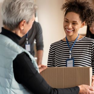 Woman smiles, hands cardboard box to another. Volunteers inside a building.