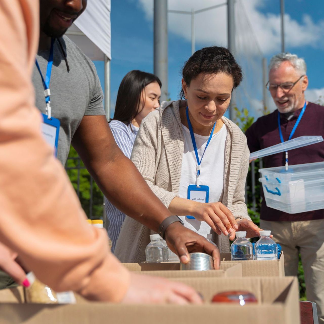 Volunteers sort donations outdoors: food, water, and supplies. People with lanyards gather near boxes and containers.