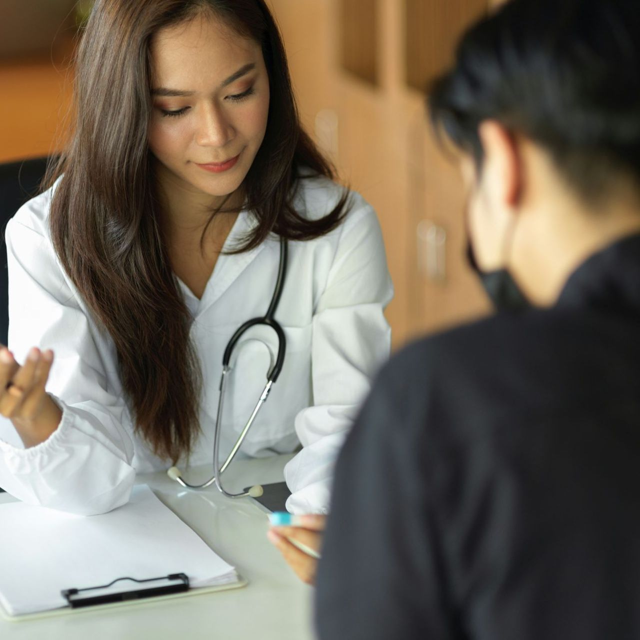 Female doctor in white coat talking to a patient at a desk; stethoscope, clipboard.