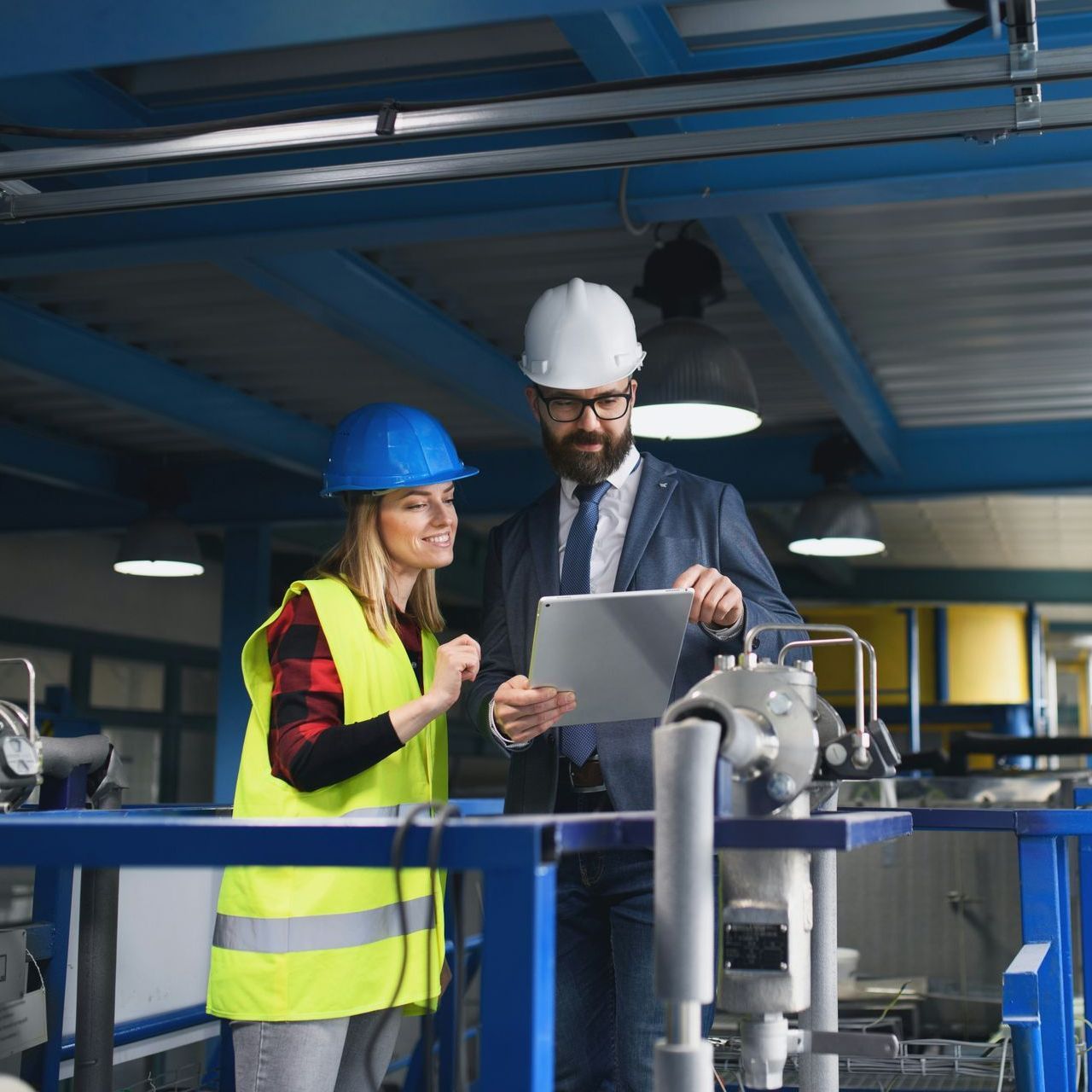 Woman in blue helmet and man in white helmet review a tablet in a factory.
