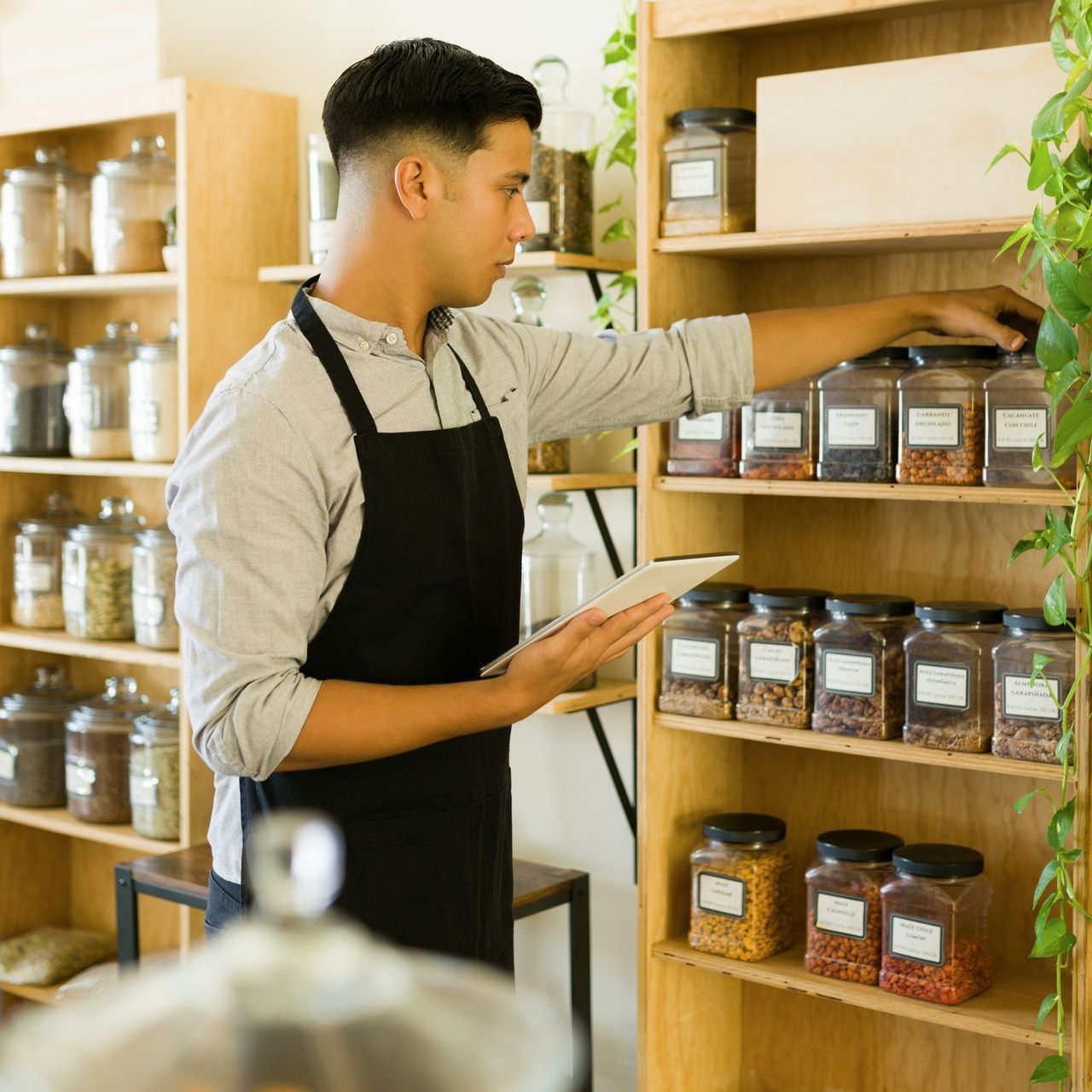 Man in apron stocking shelves, holding tablet, in a store with jars of products.