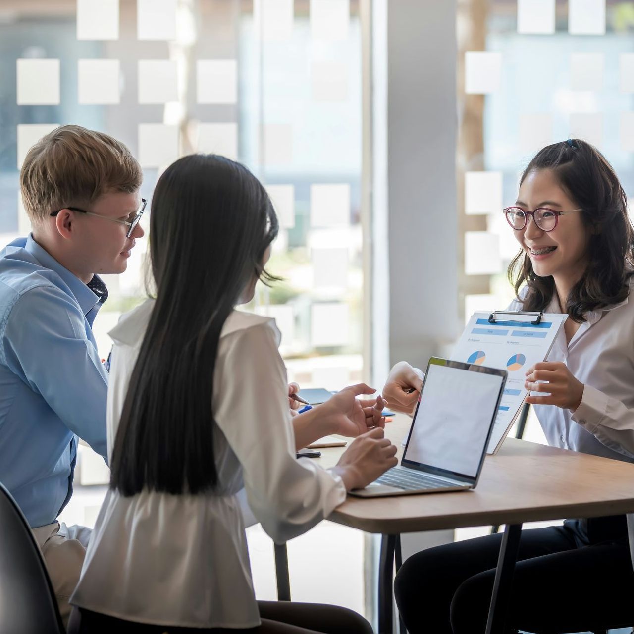 Three people at a table, discussing paperwork and a laptop. The woman at right smiles, presenting data.