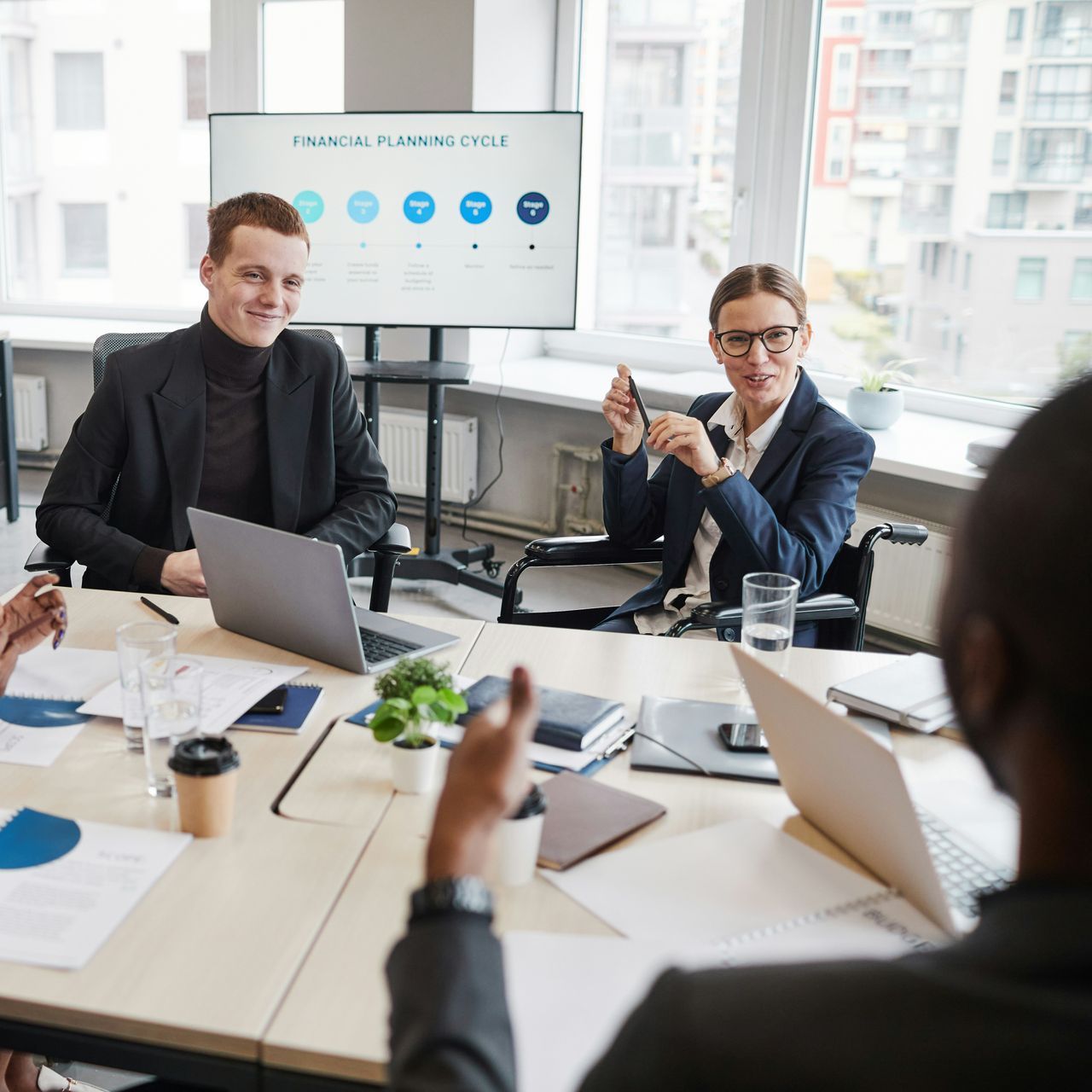 Business meeting: People around a table, with a large screen displaying charts.