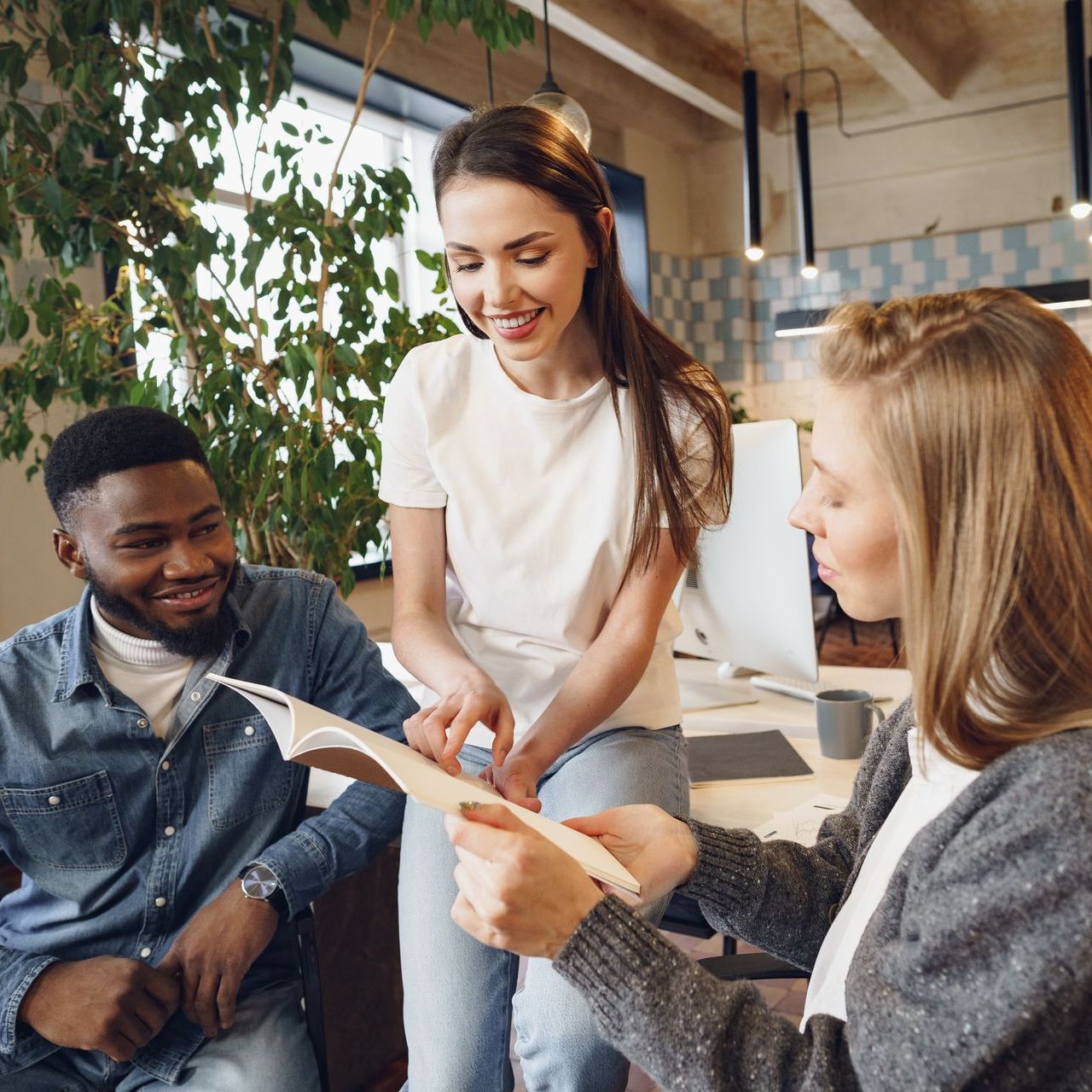 Three coworkers collaborating over papers, smiling, in a modern office with plants and light fixtures.