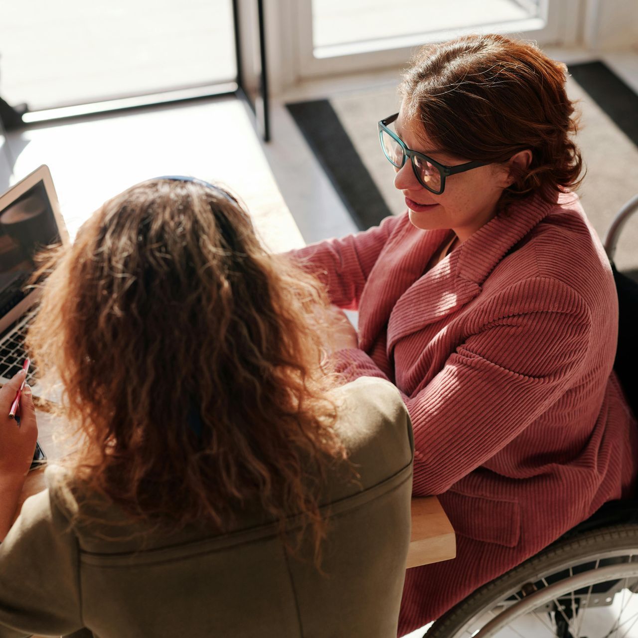 Two women at a table, one in a wheelchair. Woman in wheelchair wearing glasses, looking at laptop.