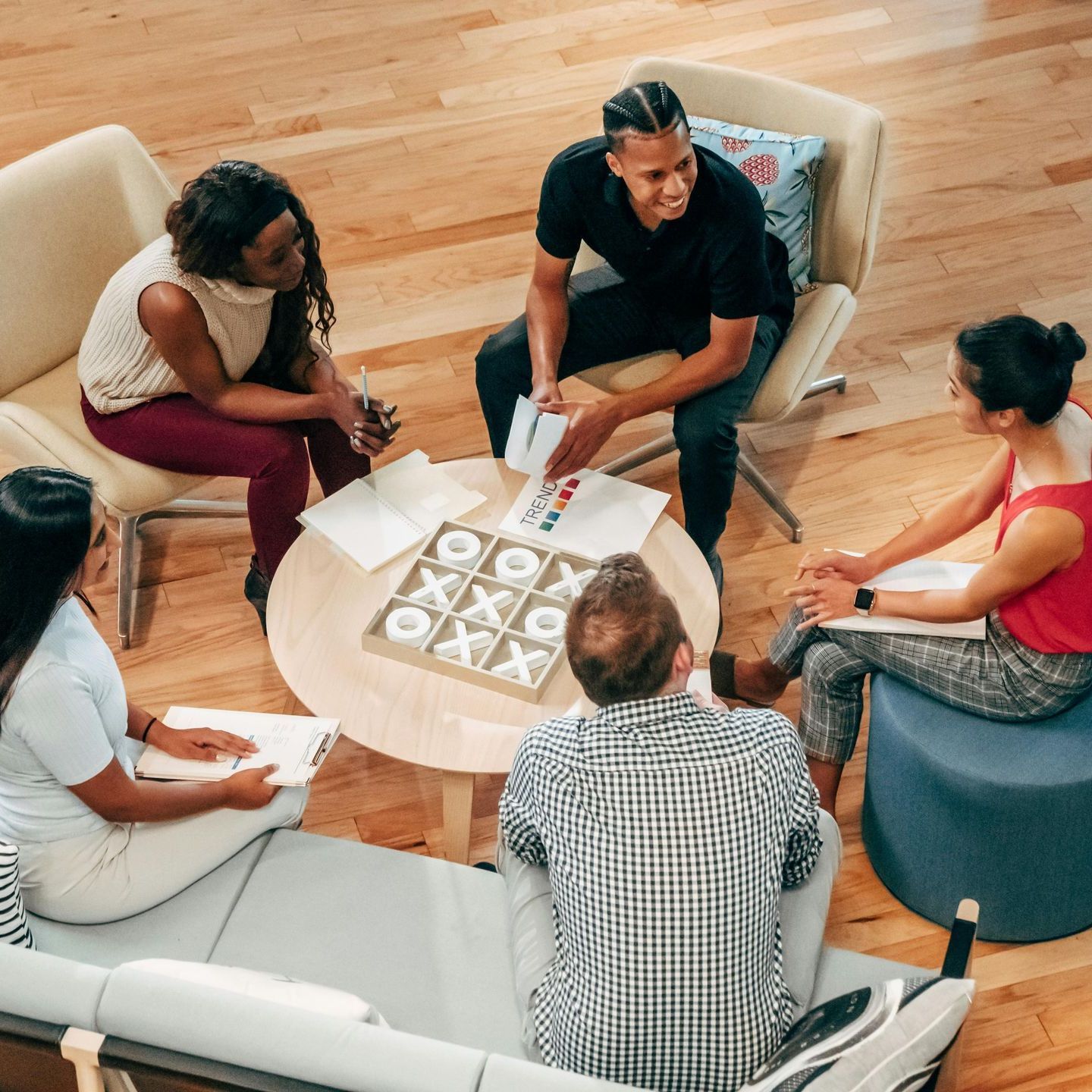 Group of diverse people in a circle, brainstorming around a table with papers and a tic-tac-toe game.