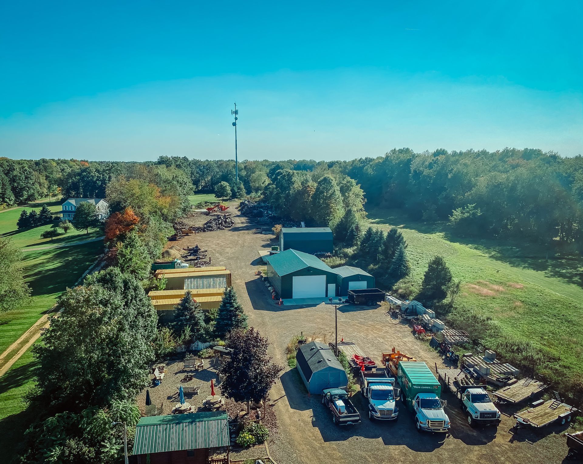 Aerial view of a business with a green roof, vehicles, and a radio tower, surrounded by trees and fields under a blue sky.