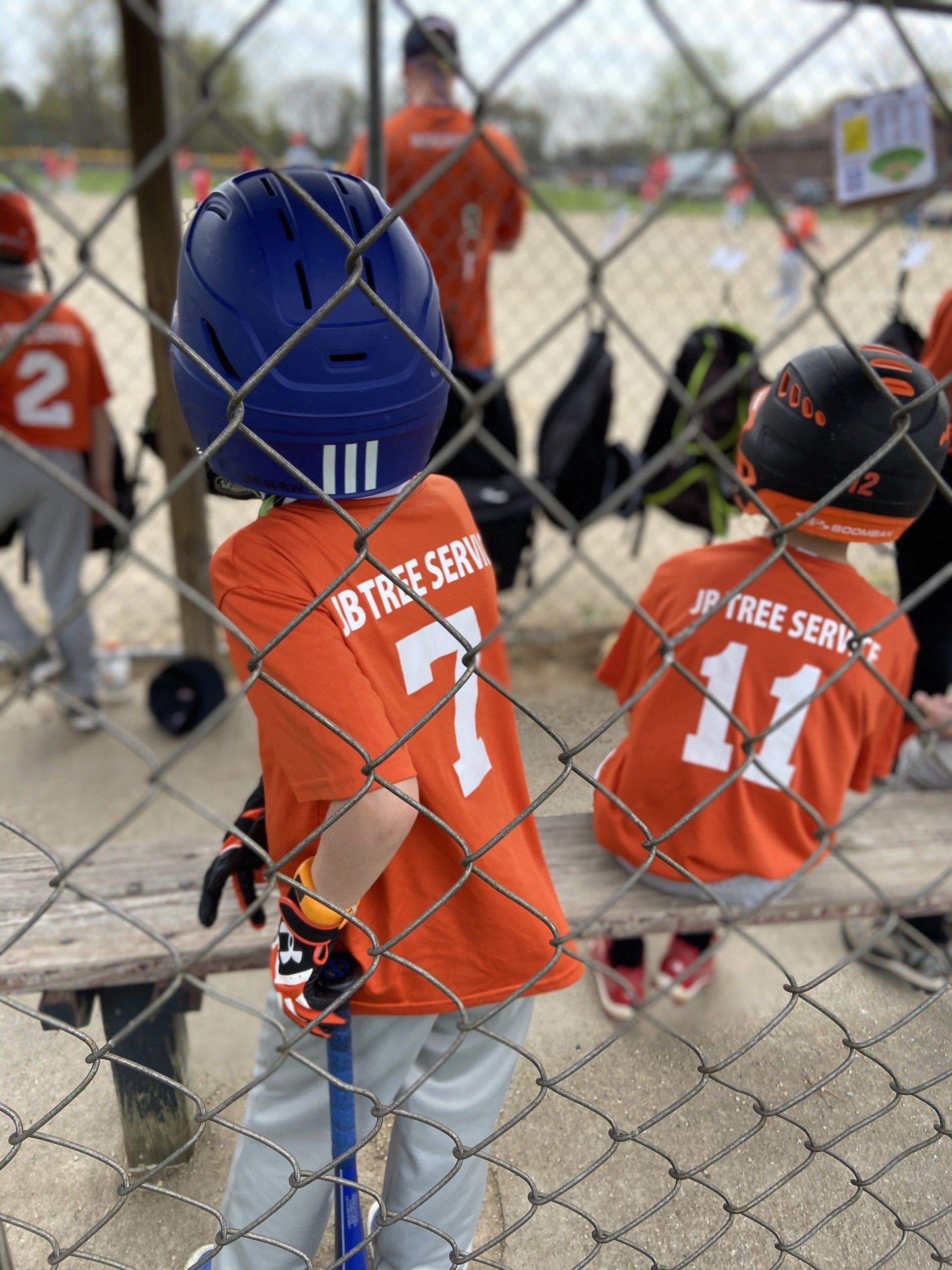 Youth baseball players in orange jerseys, helmets on, stand behind a chain-link fence, waiting.