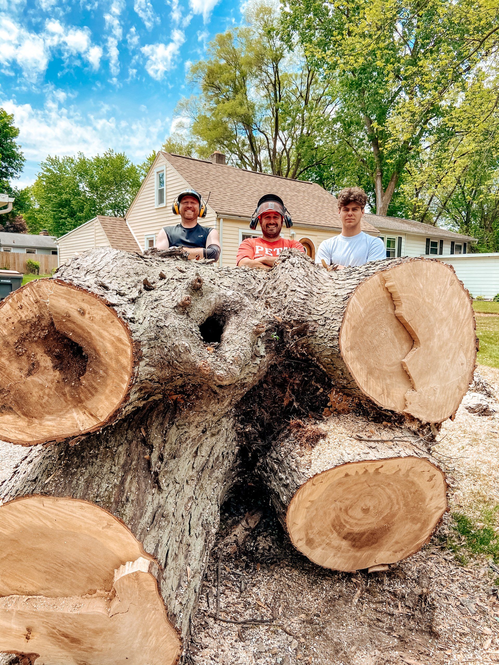 Three young men stand behind a large, cut tree trunk outdoors. They are smiling, wearing hats and safety glasses.