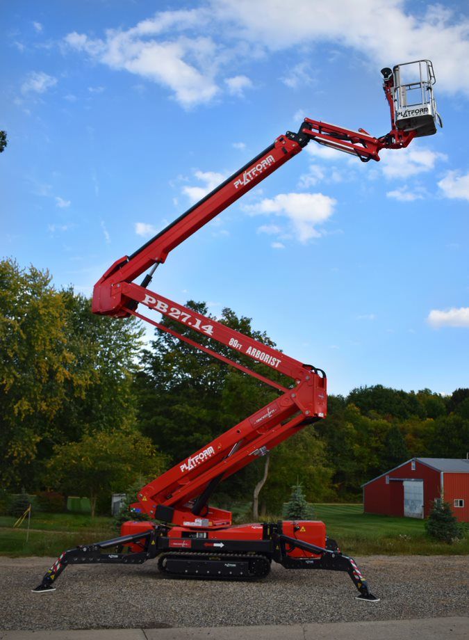 Red aerial lift with tracks and extending boom, outdoors near trees and a building.