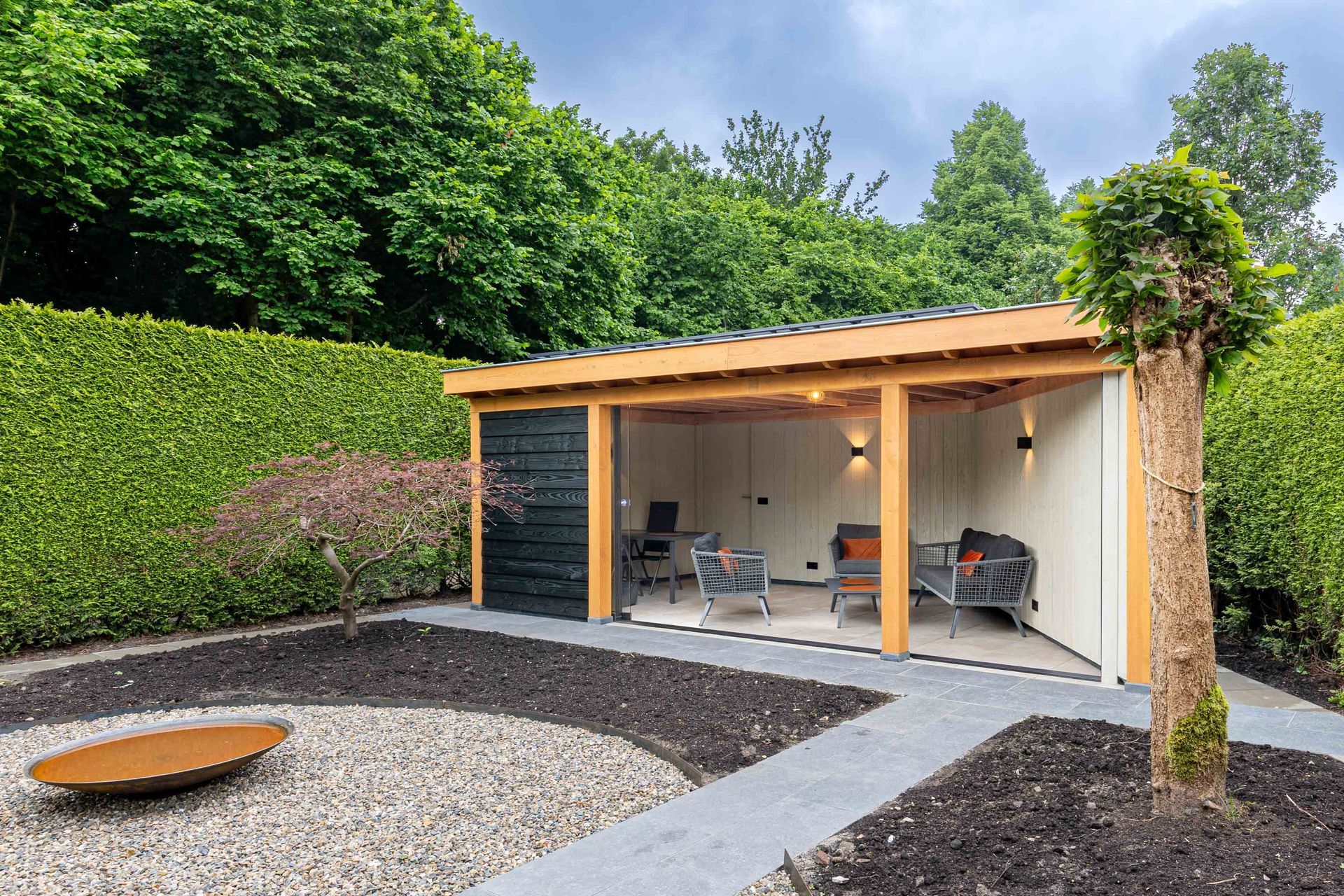 Een houten tuinpaviljoen met open voorkant en tuinmeubilair, gelegen in een achtertuin met een grindgedeelte, hagen en een boom.