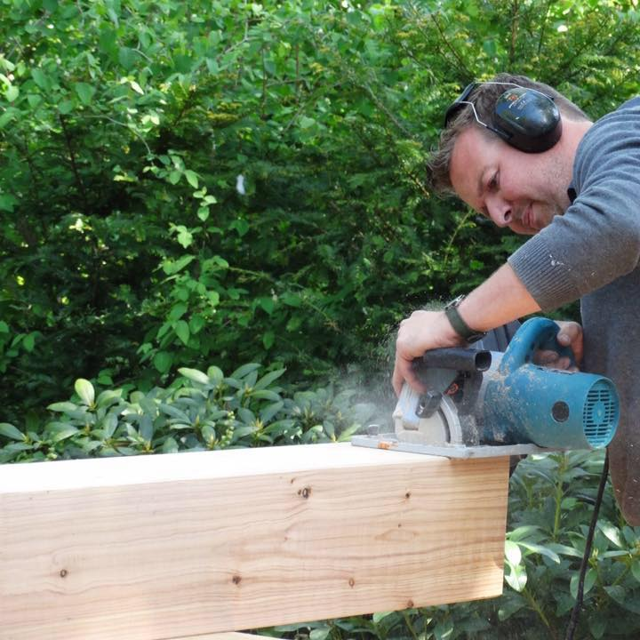 Een persoon met gehoorbescherming gebruikt een blauwe cirkelzaag om een ​​dikke houten balk door te zagen in de buitenlucht, vlakbij groen gebladerte.