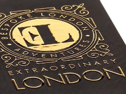 a close up of a gold logo for extraordinary london on a dark brown background.