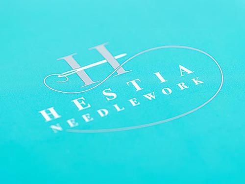 a close up of the hestia needlework logo on a blue surface.