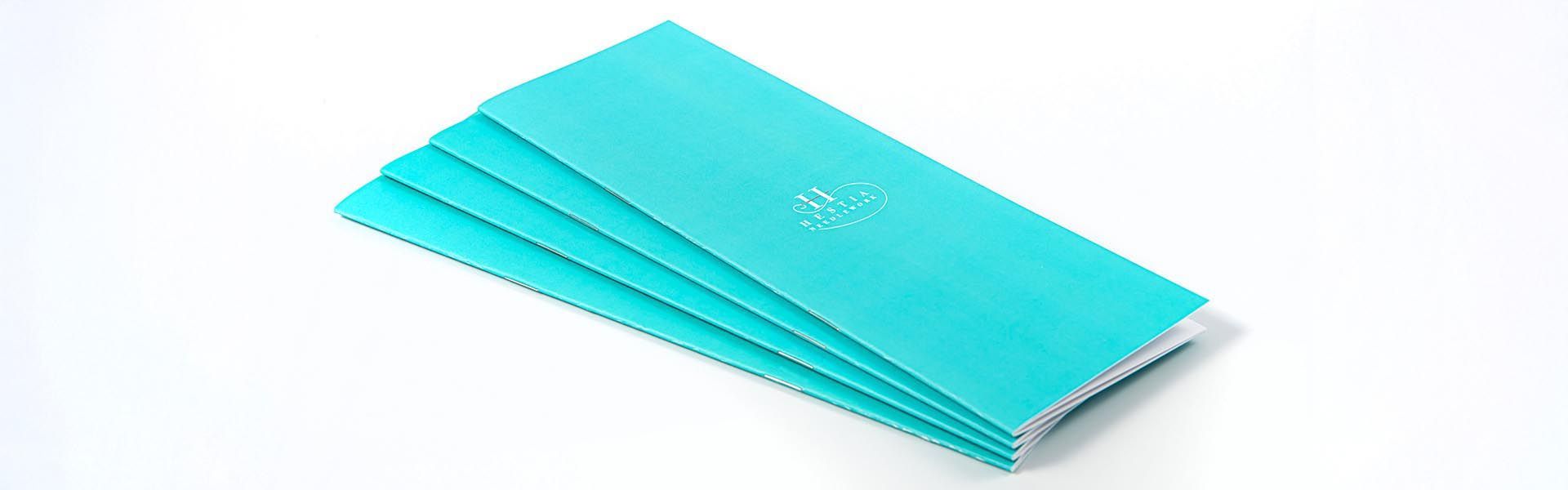 a stack of turquoise colored notebooks with the hestia needlework logo on the front