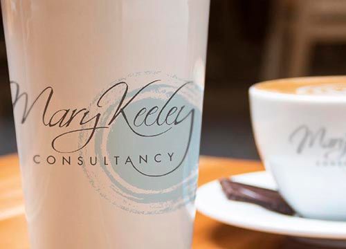 a cup that says mary keeley consultancy is on a table next to a cup of coffee .